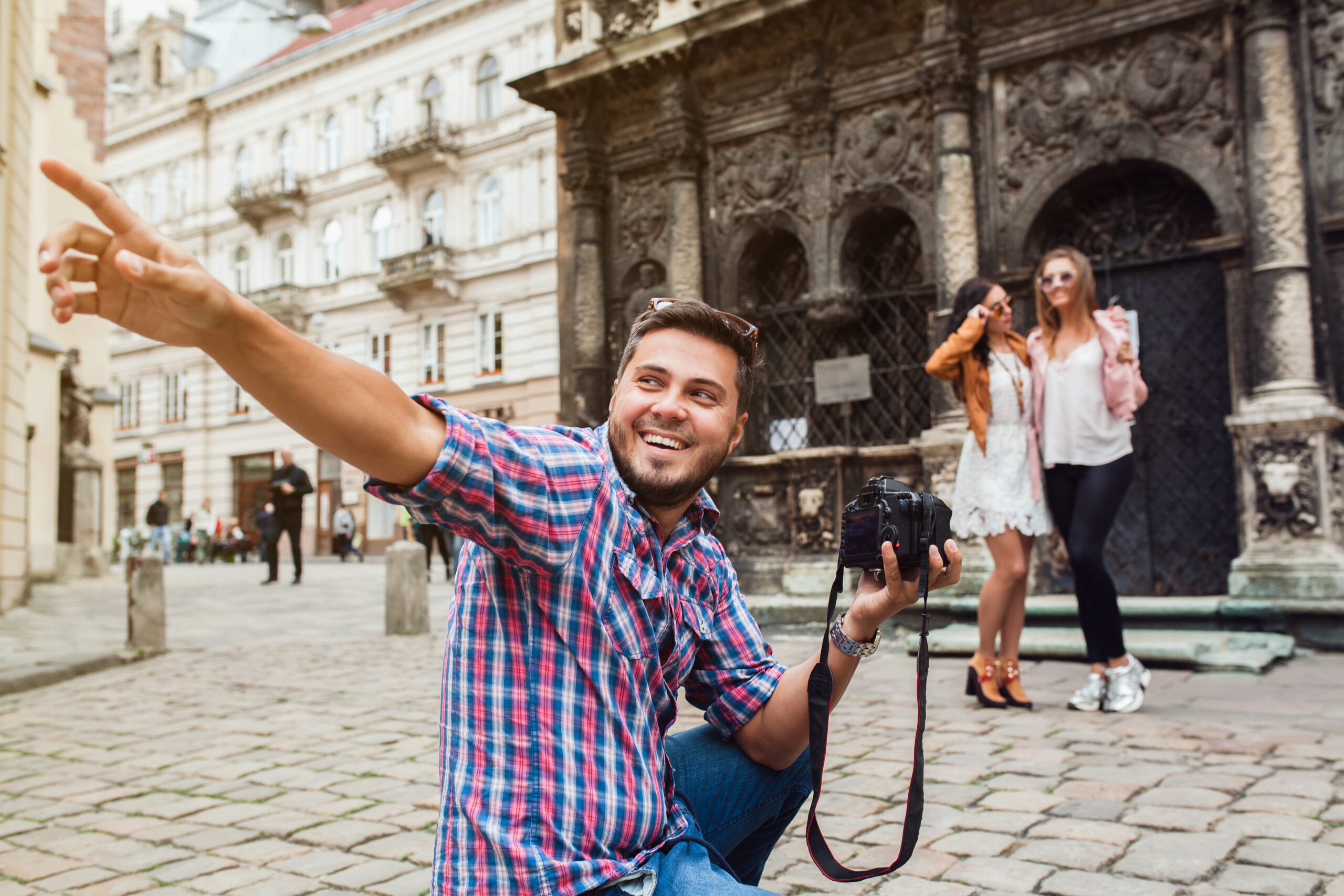 young man photographer taking pictures, holding digital photo camera, girls posing on background, friends traveling around europe, summer vacation, having fun, vintage street style