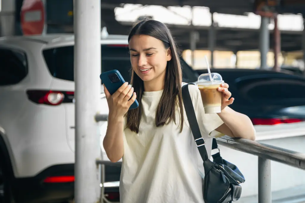 mujer joven alegre con telefono inteligente y algo en el estacionamiento