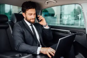 hombre de negocios guapo barbudo y sonriente trabajando en su computadora portatil y hablando por telefono movil en el asiento trasero del automovil
