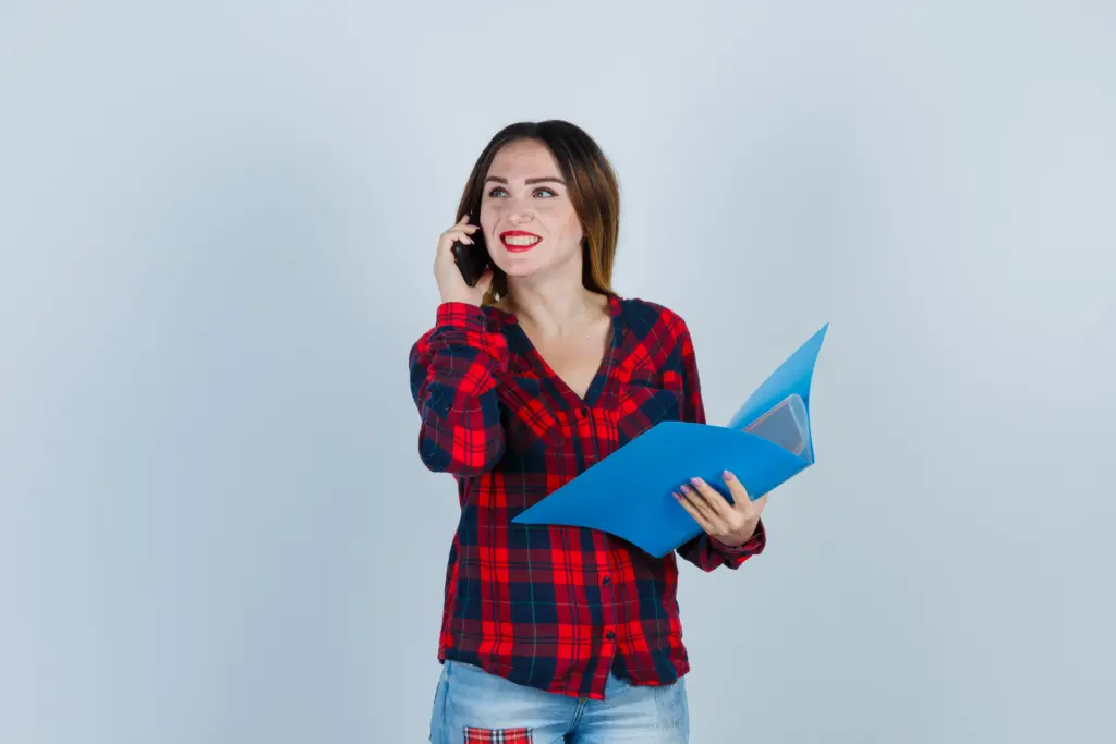 retrato de joven hermosa mujer sosteniendo la carpeta mientras habla por telefono mirando otro lado en camisa casual jeans y mirando feliz vista frontal