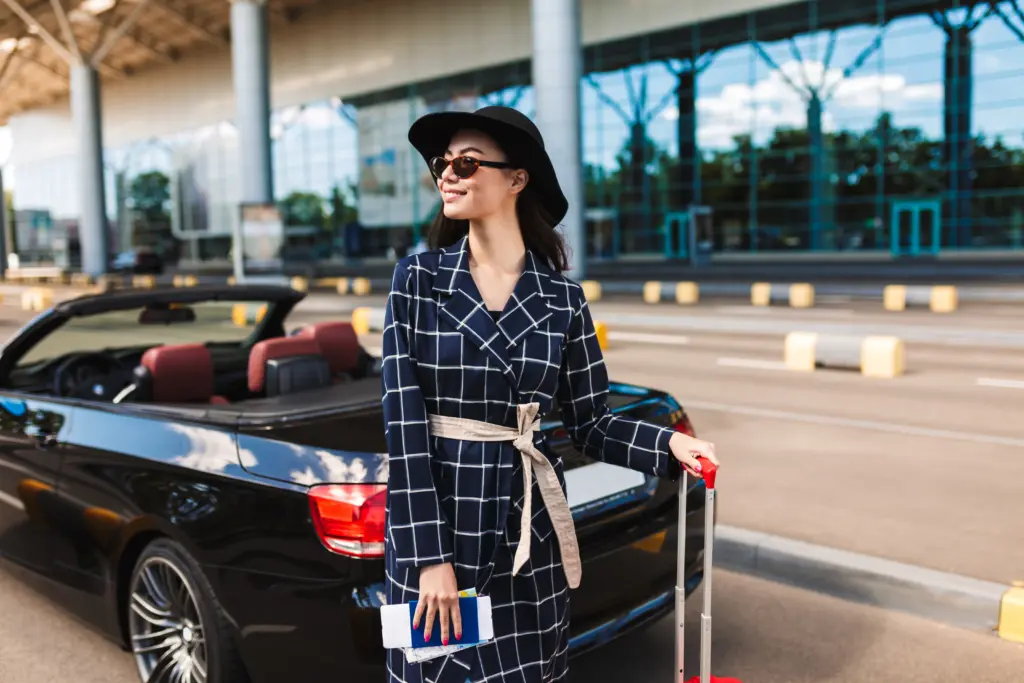 beautiful smiling girl sunglasses black hat holding passport with flight ticket joyfully looking aside near airport with cabriolet car background
