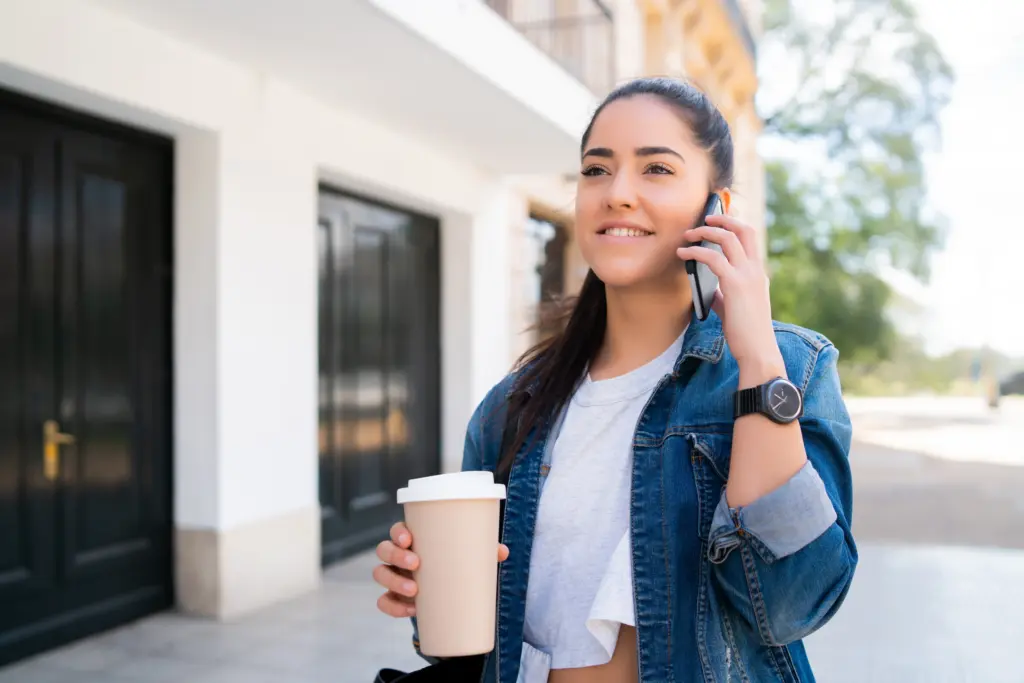 portrait young woman talking phone holding cup coffee while standing outdoors street