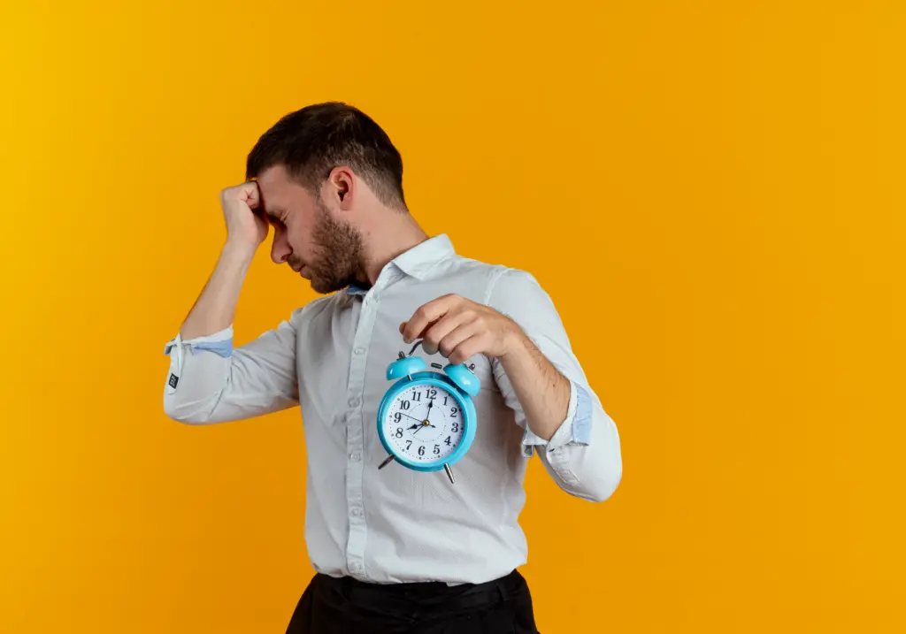 upset handsome man puts fist forehead holding alarm clock looking side isolated orange wall