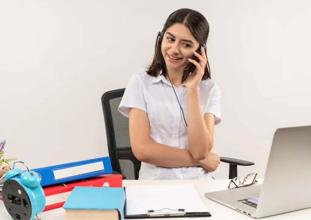 chica joven con camisa blanca y auriculares hablando por telefono movil sonriendo sentado en la mesa con carpetas y portatil sobre pared blanca