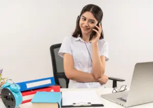 chica joven con camisa blanca y auriculares hablando por telefono movil sonriendo sentado en la mesa con carpetas y portatil sobre pared blanca