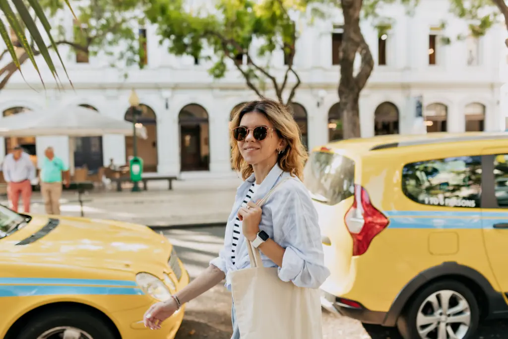 mujer increible feliz con cabello ondulado en gafas de sol con bolsa ecologica esta caminando en la soleada calle europea contra arboles exoticos y taxis de la ciudad