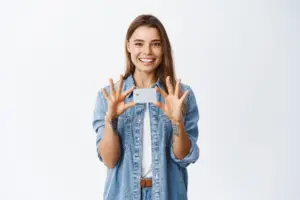 imagen de una joven mujer caucasica sonriente con emocion positiva mostrando una tarjeta de credito de plastico sobre el pecho recomendar deposito bancario de pie sobre blanco